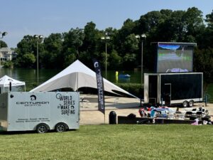 Outdoor event setup includes a trailer labeled "Centurion Boats," flags, chairs, a large tent, and a big screen near a lakeside area with trees in the background for the Wake Surf Classic. Volunteers from Centurion WSWS are on site to assist.