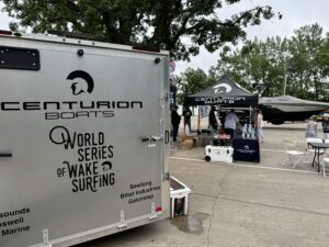 A Centurion Boats trailer and promotional tent are set up in a parking lot for the King of the Lakes Classic. People are gathered under the tent, and a wakeboarding boat is positioned nearby, showcasing Faction Marine's latest innovations.