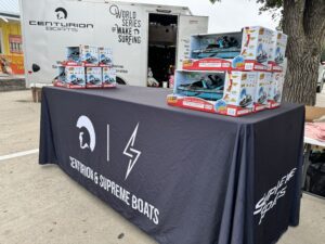 A table covered with a Centurion & Supreme Boats tablecloth displays toy boat boxes in front of a Centurion Boats trailer, proudly promoting the King of the Lakes event at the World Series of Wake Surfing.