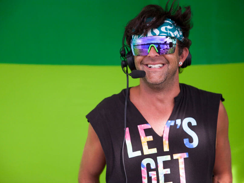 A man wearing sunglasses and a t-shirt stands confidently in front of a green background, captivating the attention of onlookers who are both competitors and fans.
