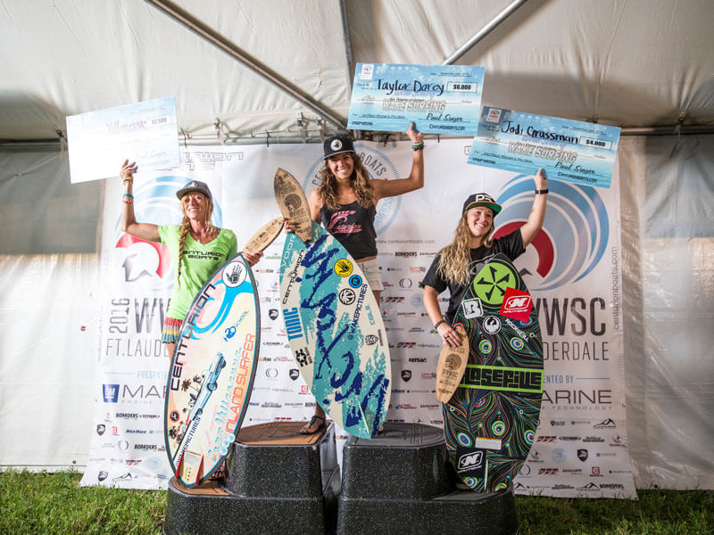 Three female surfers standing on top of a tent with their surfboards during a wake surfing competition.