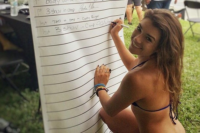 A woman writing on a white board while wearing a bikini, captivating surfers and fans.