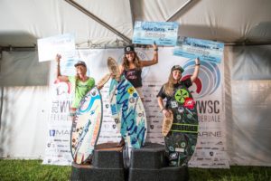 Three surfers standing on top of a tent with their surfboards, participating in a contest.