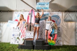 Three competitors standing on top of a stage with surfboards.