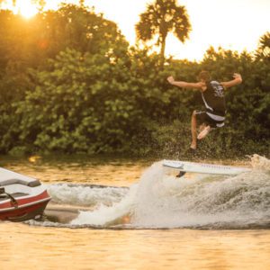 Two competitors in a wake surfing contest, with one man on a surfboard jumping over a boat.