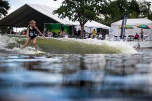 A contestant is wake surfing on a surfboard in front of a tent during a competition.