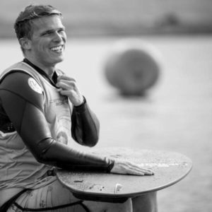 A man in a wetsuit smiling while holding a surfboard, ready to impress the fans at a surfing contest.