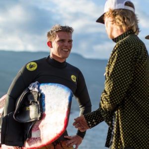 Two men in wetsuits shaking hands while holding surfboards at a wake surfing contest.