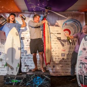 Three competitors standing on a contest podium with their surfboards.