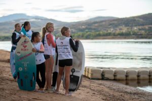 A group of competitors standing on a beach with surfboards, ready for wake surfing.