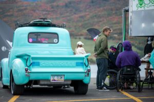A turquoise truck with a man in a wheelchair standing next to it, preparing for a wake surfing contest as enthusiastic fans cheer him on.