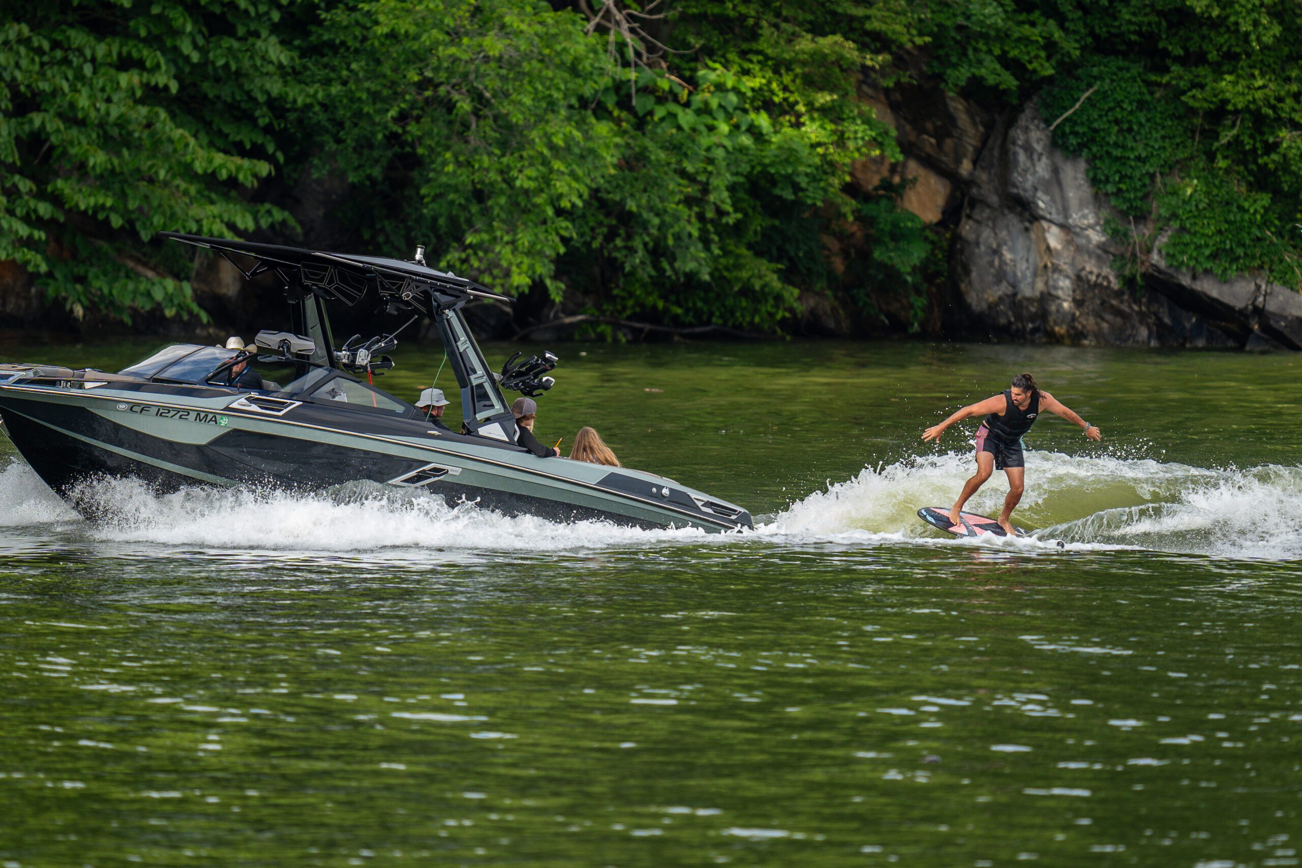 A boat tows a person wakeboarding on calm green waters surrounded by lush greenery, as part of the Volunteer Wake Surf Classic hosted by CBK Watersports.