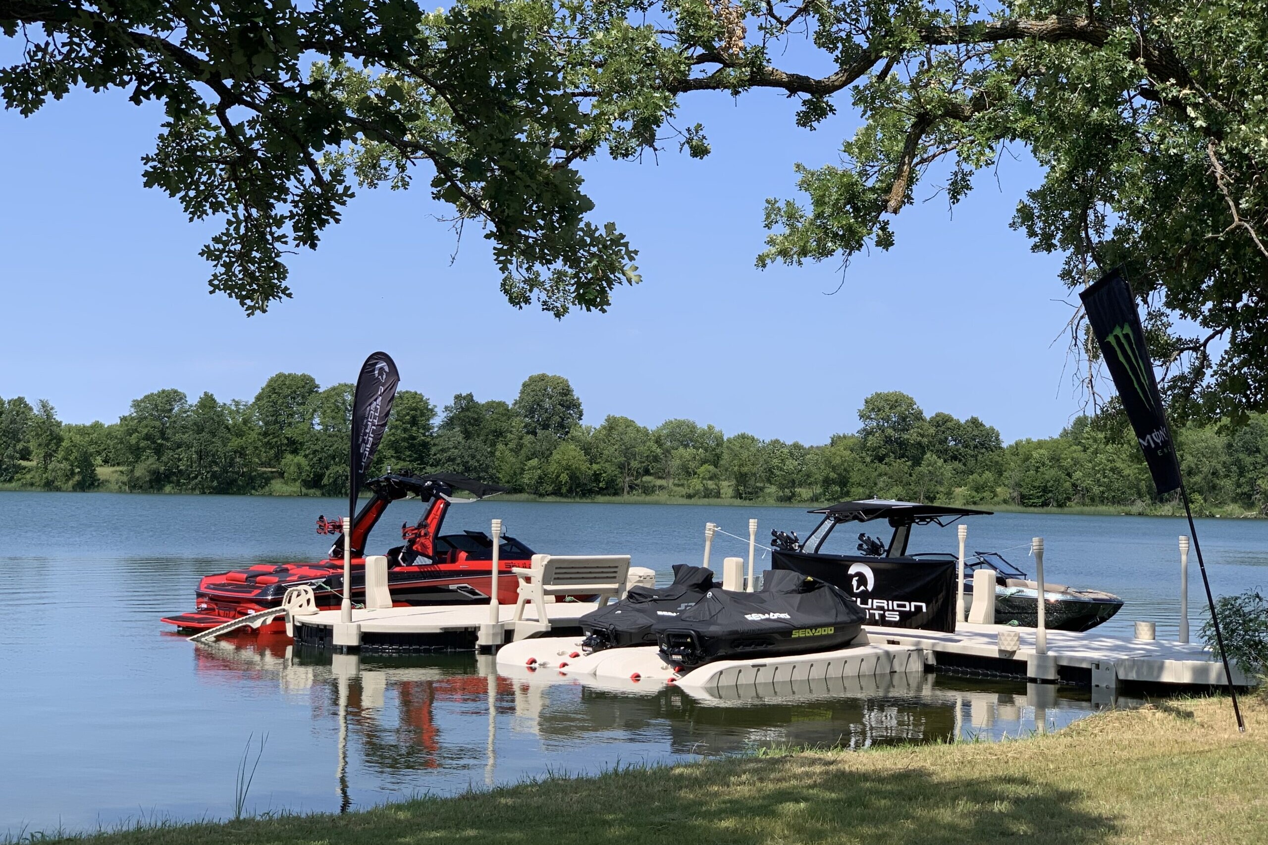 A lakeside scene with several boats and jet skis docked at a pier, some preparing for the King of the Lakes Classic. Trees frame the water, and flags from Surf MN and Faction Marine are visible near the boats under a clear, blue sky.