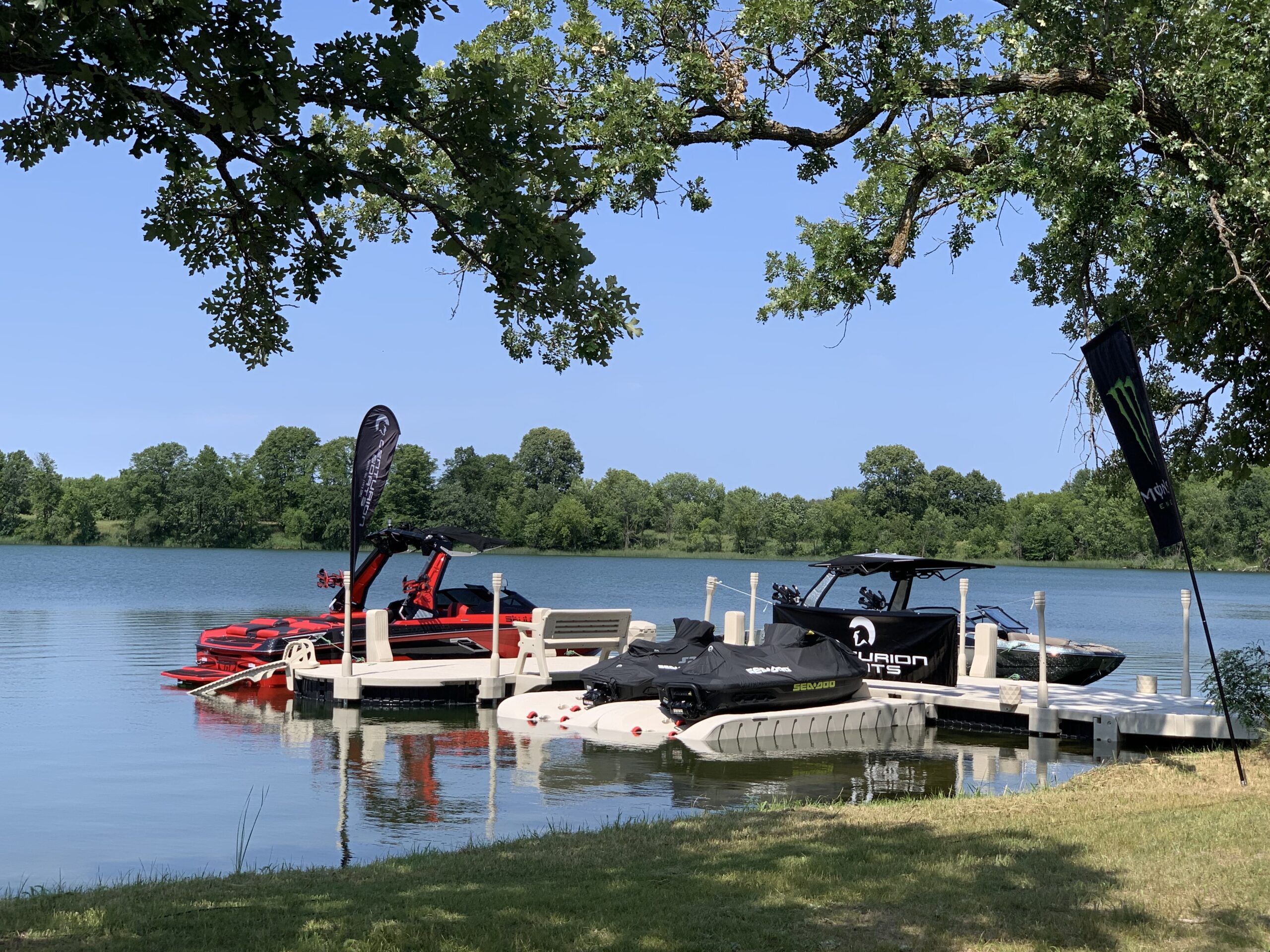 A lakeside scene with several boats and jet skis docked at a pier, some preparing for the King of the Lakes Classic. Trees frame the water, and flags from Surf MN and Faction Marine are visible near the boats under a clear, blue sky.