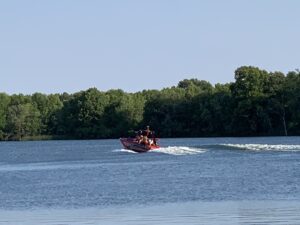 A red motorboat with several people on board, sporting the Faction Marine logo, glides across a calm lake with trees lining the shore under a clear sky.