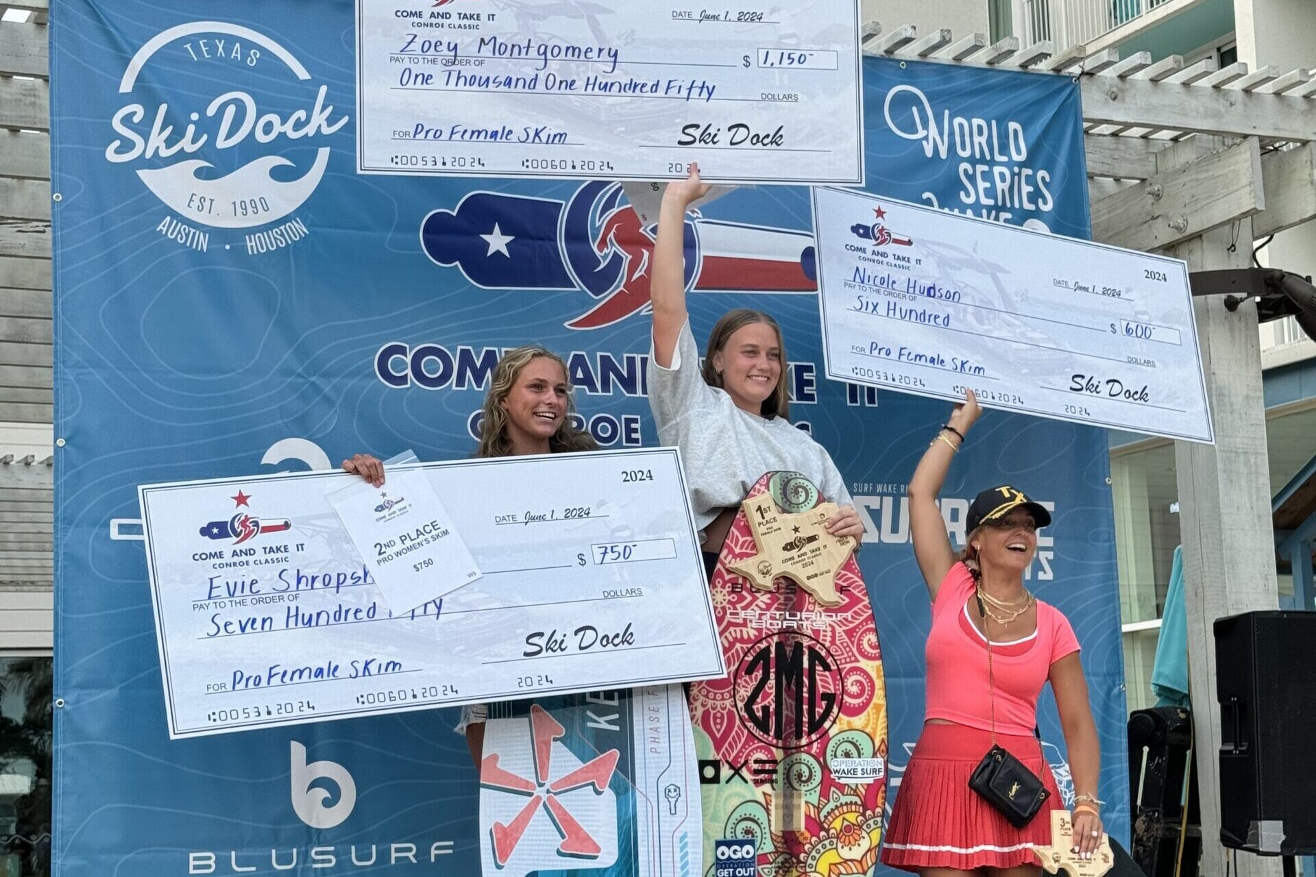 Three women pose on a winners' podium during the Conroe Classic Day 2 Finals, holding oversized prize checks and small awards, with one woman clutching a trophy. The backdrop displays logos and promotional signage from sponsors, including Centurion WSWS.
