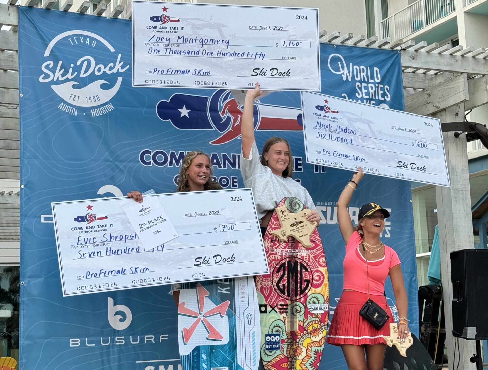 Three women pose on a winners' podium during the Conroe Classic Day 2 Finals, holding oversized prize checks and small awards, with one woman clutching a trophy. The backdrop displays logos and promotional signage from sponsors, including Centurion WSWS.