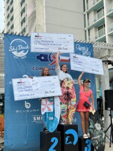 Three women stand on podiums holding large checks and trophies at the Conroe Classic surfing competition. The first place is in the center, with the second on the left and the third on the right, celebrating their achievements during Day 2 Finals of the Centurion WSWS.