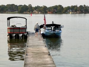 A person walks barefoot on a wooden ski dock towards a pontoon boat and a small motorboat docked on a calm lake. An American flag is mounted on the motorboat, possibly enjoying the annual Conroe Classic. Trees and houses are visible in the background.
