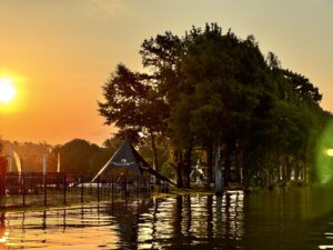 A sun sets over a lakeside scene, casting a warm glow. Trees and a tent are visible by the water, with a few people near the tent. A fence runs along the water's edge, adding to the tranquil vibe of this Centurion WSWS event.