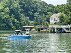 A blue boat with passengers cruises on a lake near a large house with a waterfront deck, surrounded by lush green trees. Nearby, CBK Watersports enthusiasts recall the excitement of the Centurion WSWS Volunteer Wake Surf Classic's Day 1 Recap.