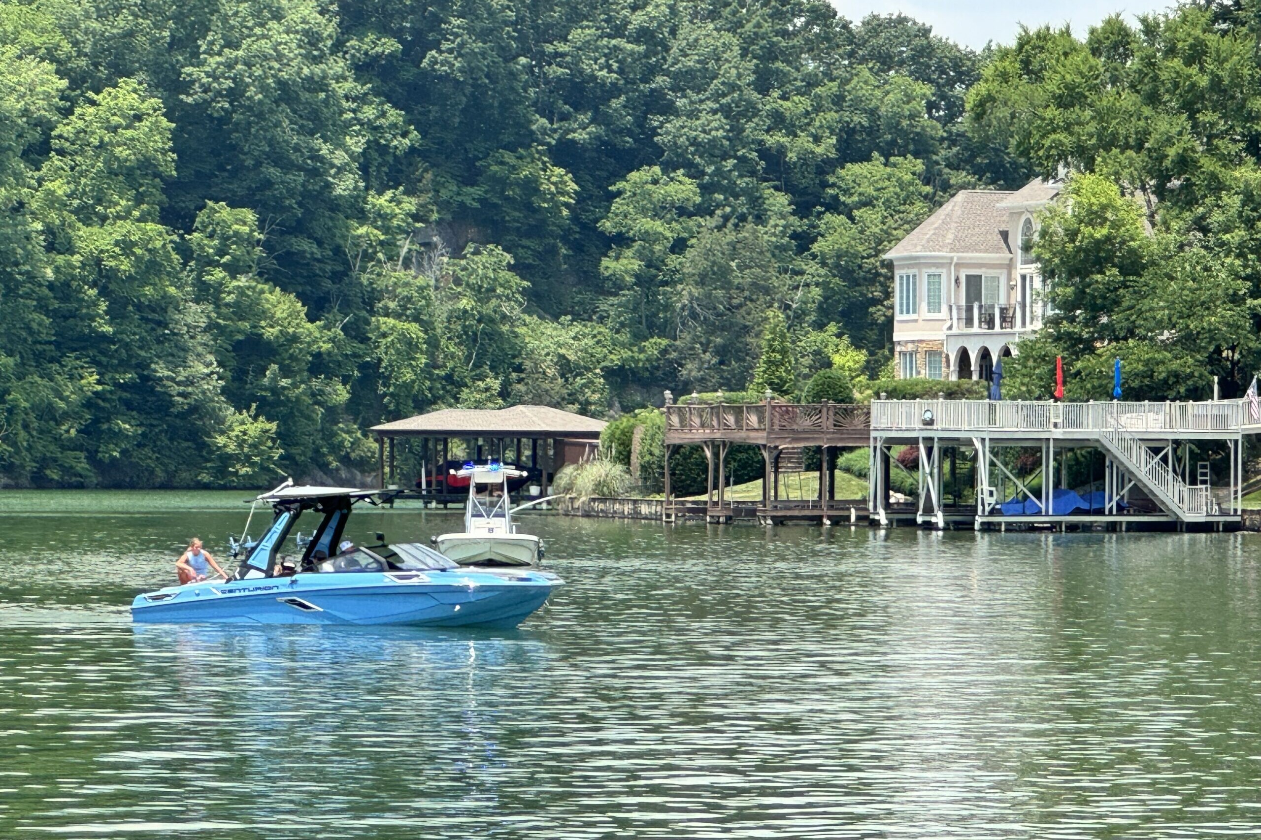 A blue boat with passengers cruises on a lake near a large house with a waterfront deck, surrounded by lush green trees. Nearby, CBK Watersports enthusiasts recall the excitement of the Centurion WSWS Volunteer Wake Surf Classic's Day 1 Recap.