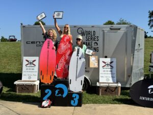 Three individuals stand on podiums labeled 1, 2, and 3, holding certificates and surfboards. A trailer behind them displays "2024 World Series Wake Surf." Various banners and signs add a classic touch to the scene.