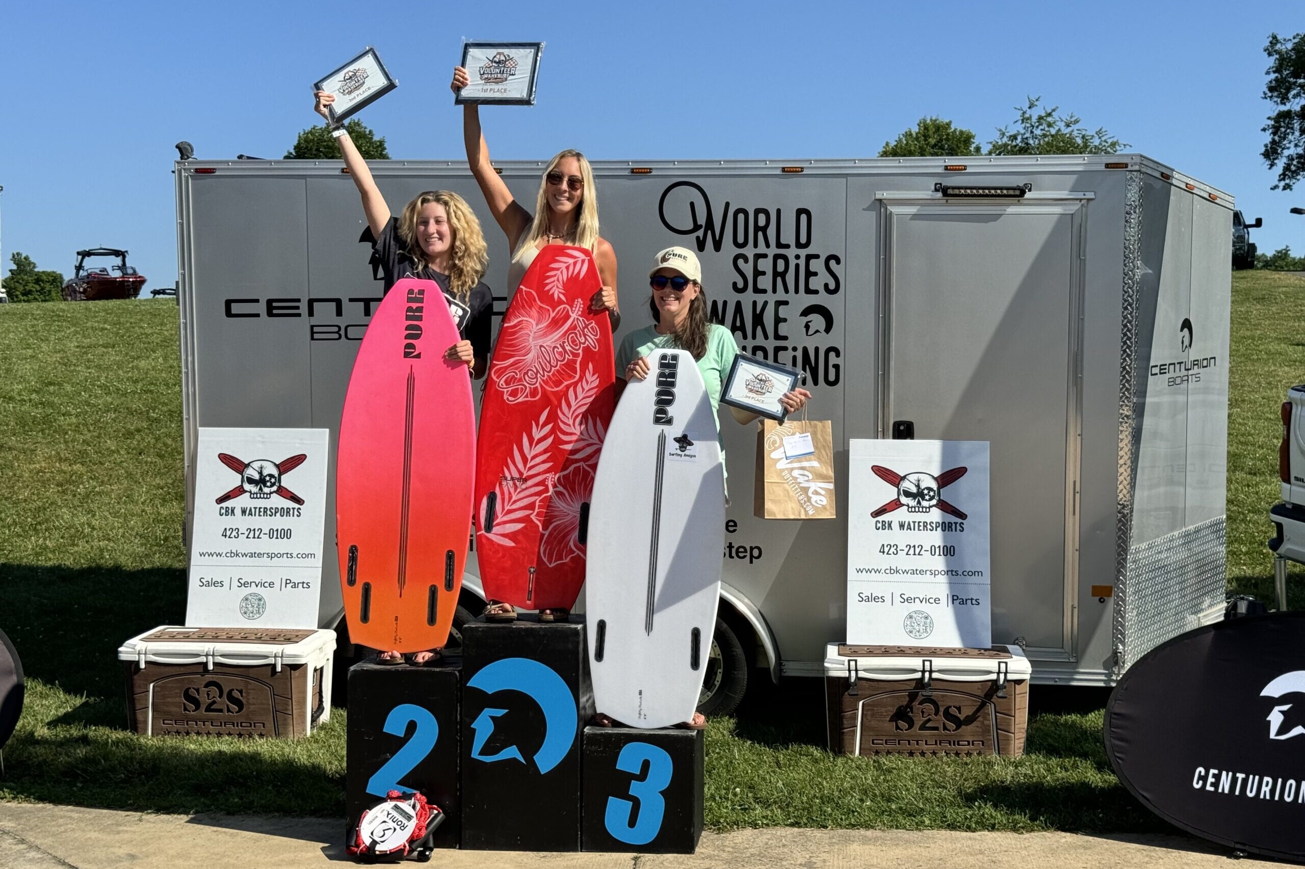 Three individuals stand on podiums labeled 1, 2, and 3, holding certificates and surfboards. A trailer behind them displays 