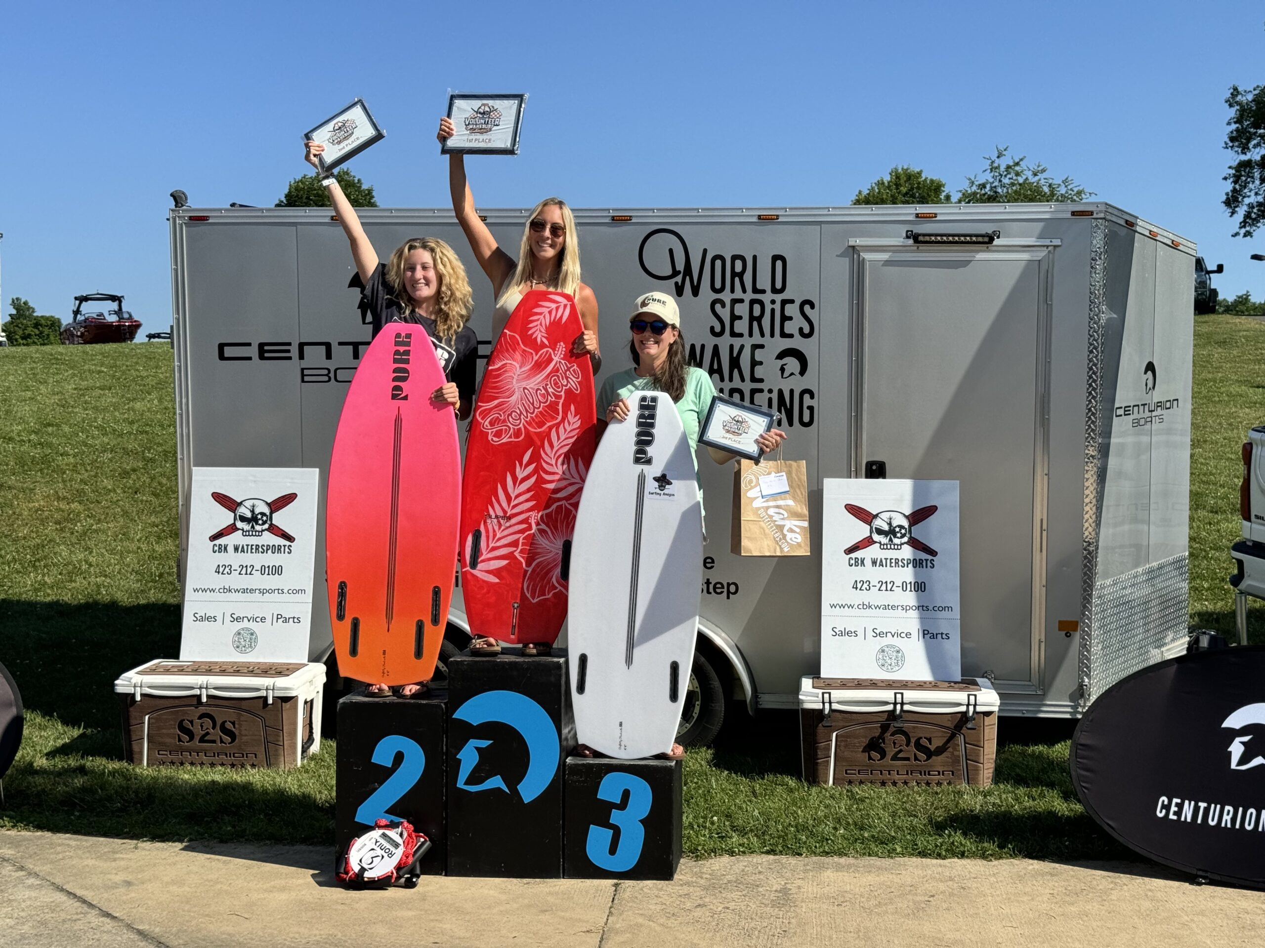 Three individuals stand on podiums labeled 1, 2, and 3, holding certificates and surfboards. A trailer behind them displays 