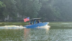 A blue Centurion WSWS motorboat with an American flag travels on a calm body of water, with a wooded shoreline in the background.