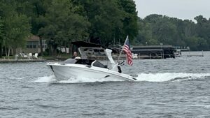 A white motorboat with an American flag cruises on a lake surrounded by green trees and lakeside houses.