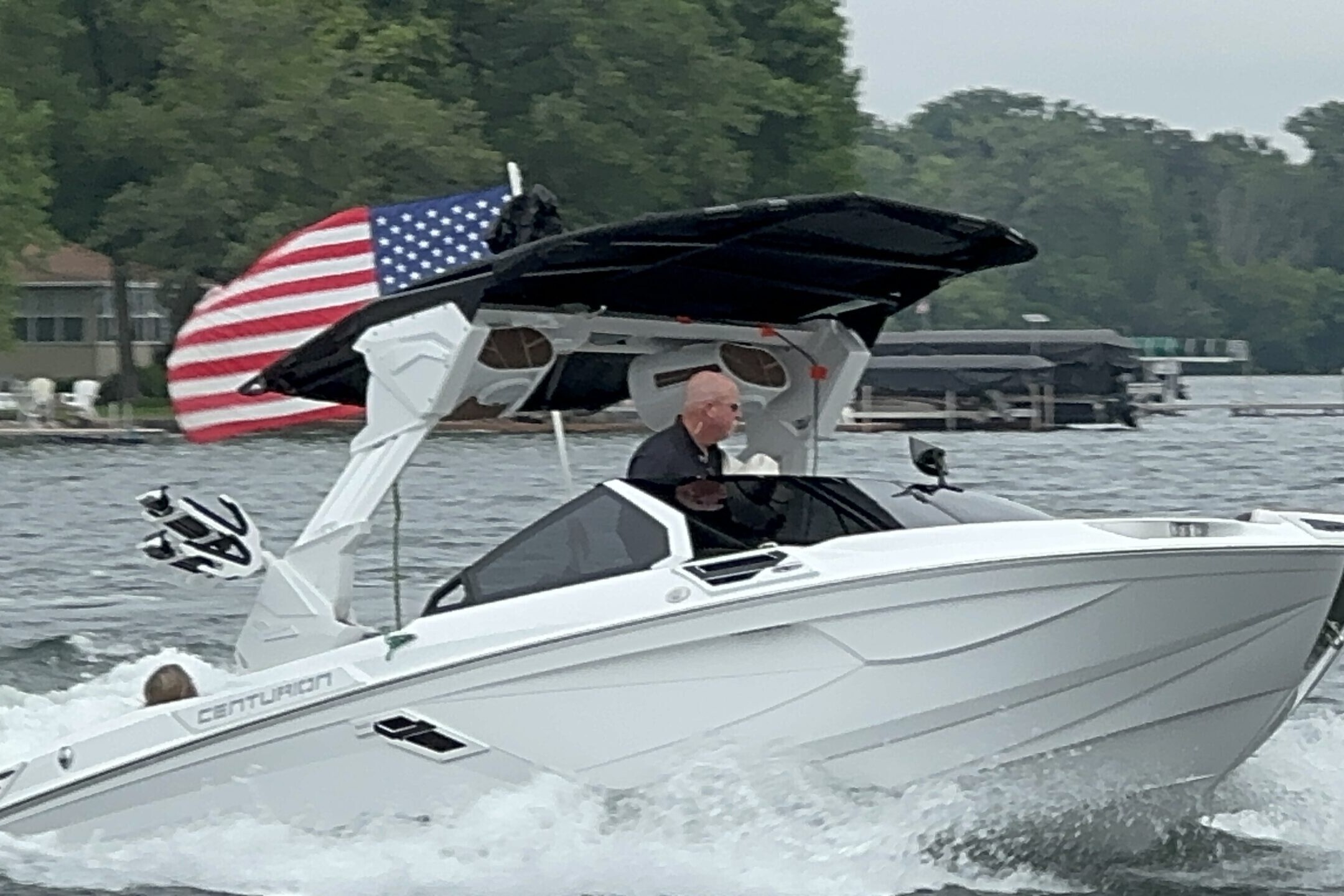 A person navigates a white Centurion boat on a lake, with an American flag displayed on the boat's canopy. Trees and houses are visible in the background, showcasing the serene setting of the King of the Lakes Classic.