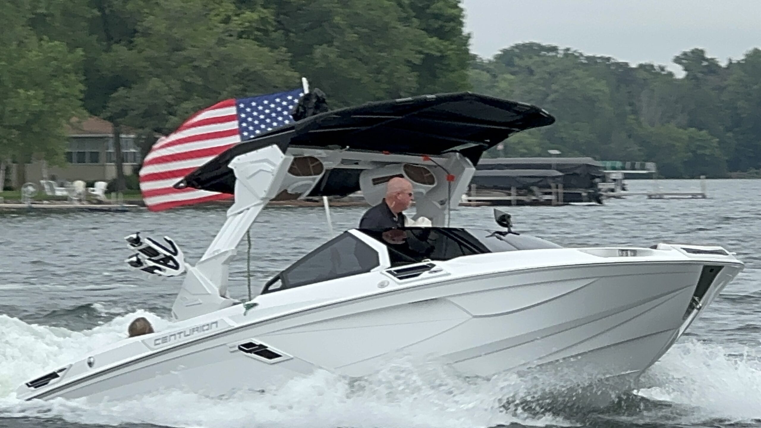 A person navigates a white Centurion boat on a lake, with an American flag displayed on the boat's canopy. Trees and houses are visible in the background, showcasing the serene setting of the King of the Lakes Classic.