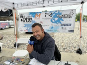 Man holding a microphone, sitting under a tented booth at the King of the Lakes Classic outdoor event. Various sponsor banners, including Surf MN and Faction Marine, are displayed behind him.