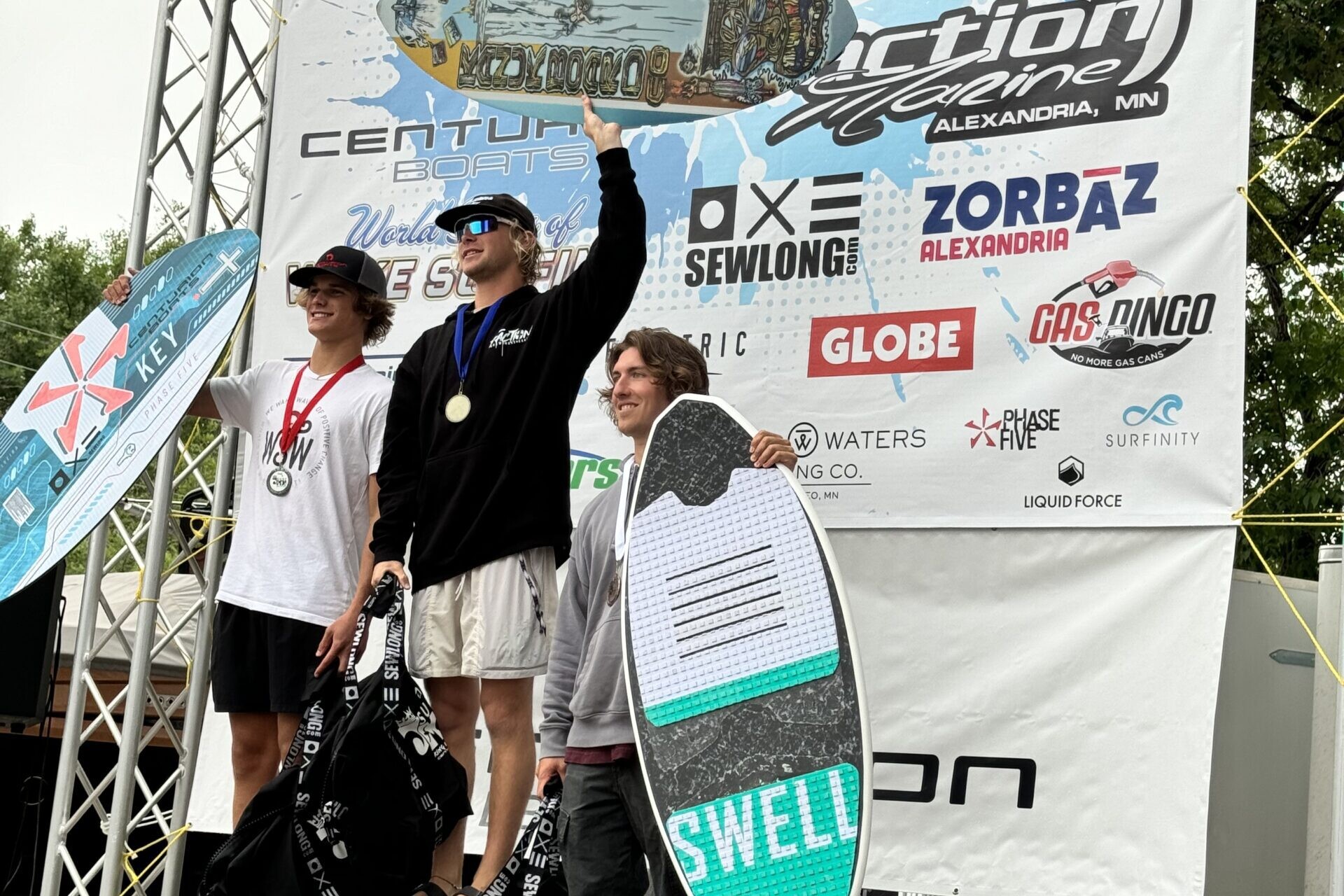 Three individuals stand on the winners' podium at a surf competition, with medals around their necks and surfboards behind them. The backdrop displays the competition's name, 