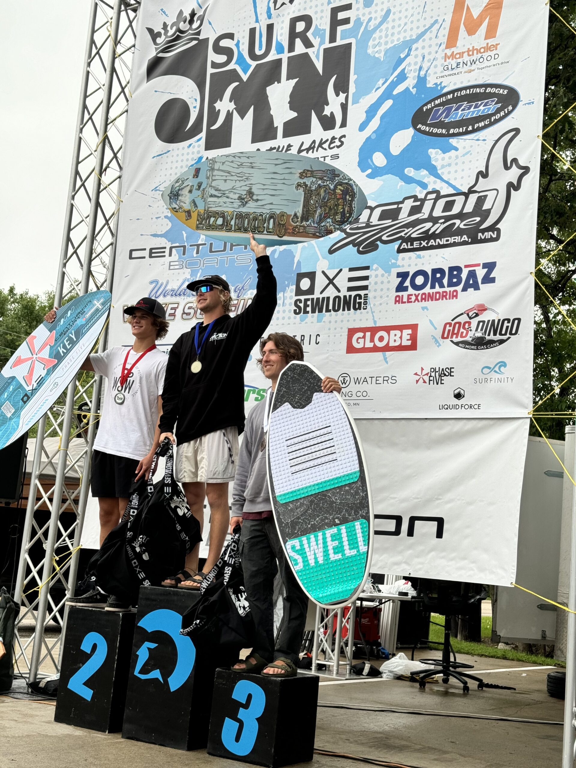 Three individuals stand on the winners' podium at a surf competition, with medals around their necks and surfboards behind them. The backdrop displays the competition's name, 