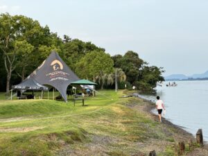 People walking by a lakeside with a tent setup labeled "CAMP MASTER" and tables underneath. Trees surround the area, with a distant view of a Centurion Wake Surf boat on the water.