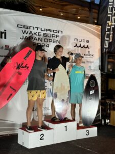 Three surfers stand on a podium holding surfboards and trophies at the Centurion Wake Surf Japan Open 2024 WSWS. The person in the middle is in first place, the person on the left is in second, and the person on the right is in third.