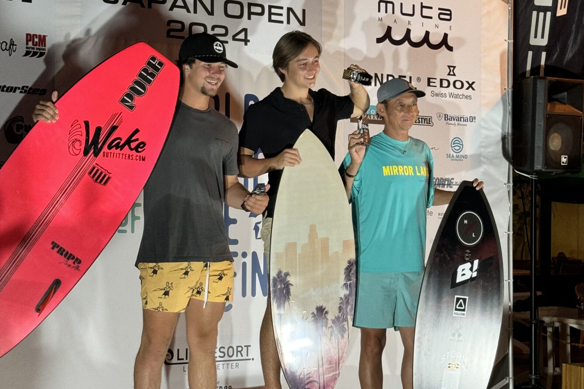 Three surfers stand on a podium holding surfboards and trophies at the Centurion Wake Surf Japan Open 2024 WSWS. The person in the middle is in first place, the person on the left is in second, and the person on the right is in third.