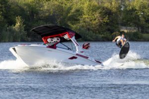 A person is wakeboarding behind a white motorboat on a lake, surrounded by trees, offering a perfect recap of a thrilling session reminiscent of the 2024 World Wake Surfing Championship.