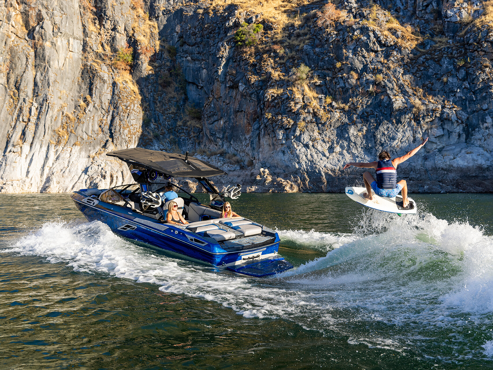 A person wakeboards on a wave created by The Centurion Ri Series, a blue and black boat expertly navigating a rocky canyon waterway.