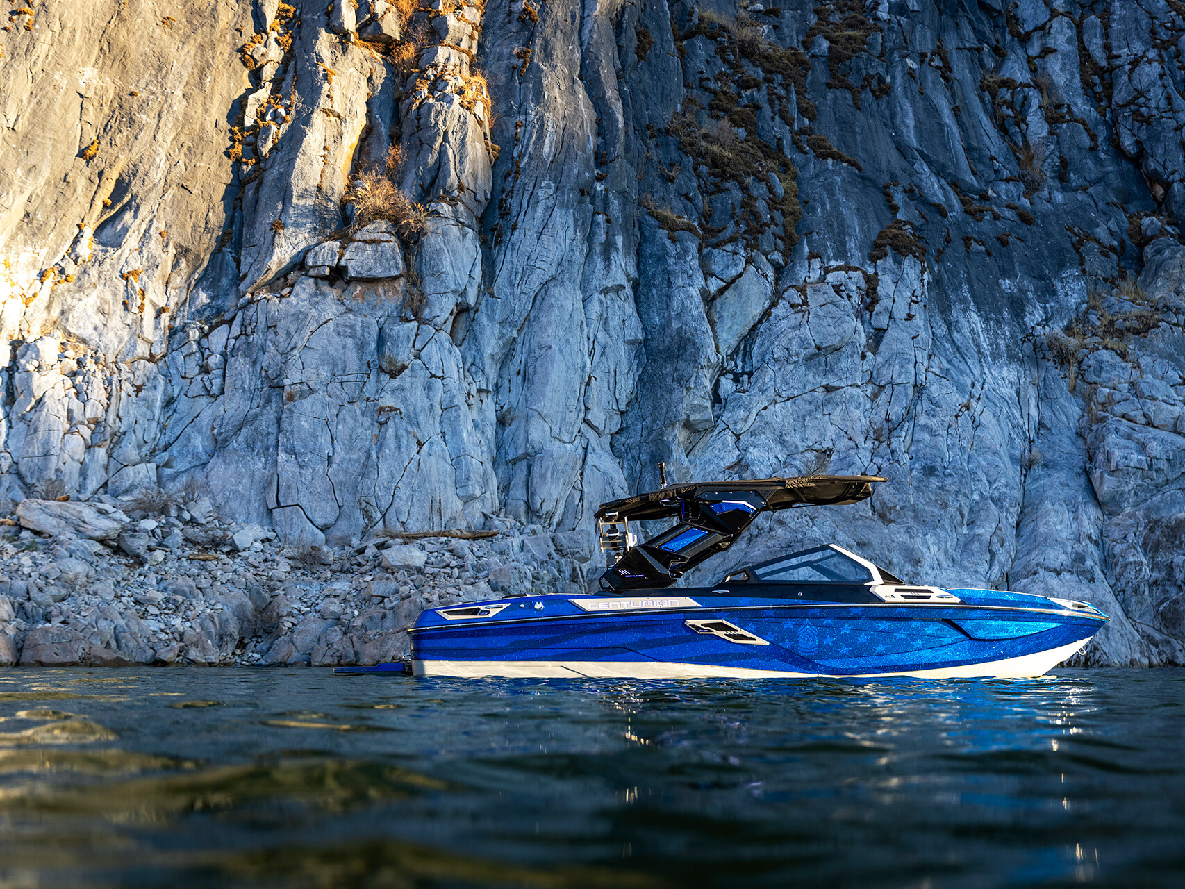 The Centurion Ri Seris, a blue boat, floats on water near a tall, rocky cliff under natural light.