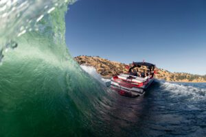 The Centurion Ri Seris navigates a large wave on a sunny day, with several passengers onboard and dry hills in the background.