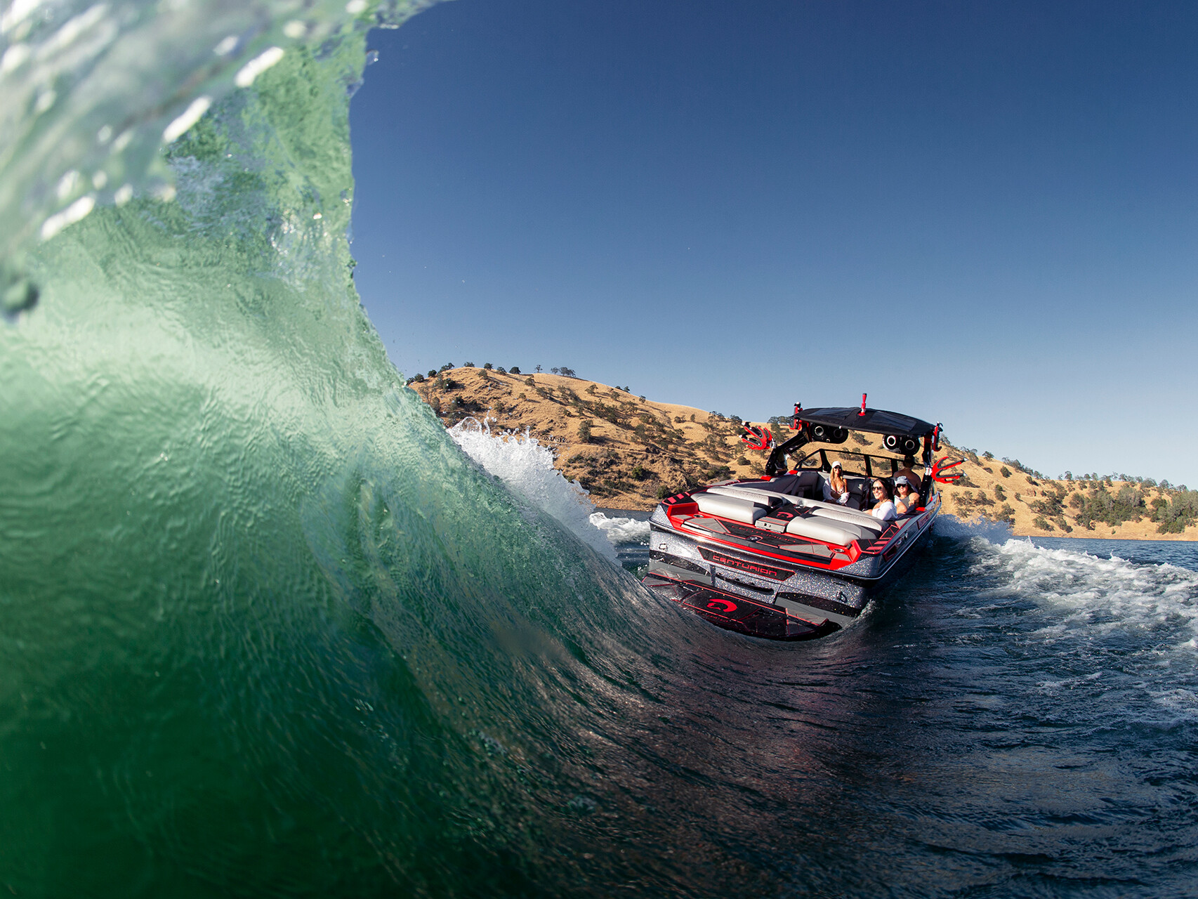 The Centurion Ri Seris navigates a large wave on a sunny day, with several passengers onboard and dry hills in the background.