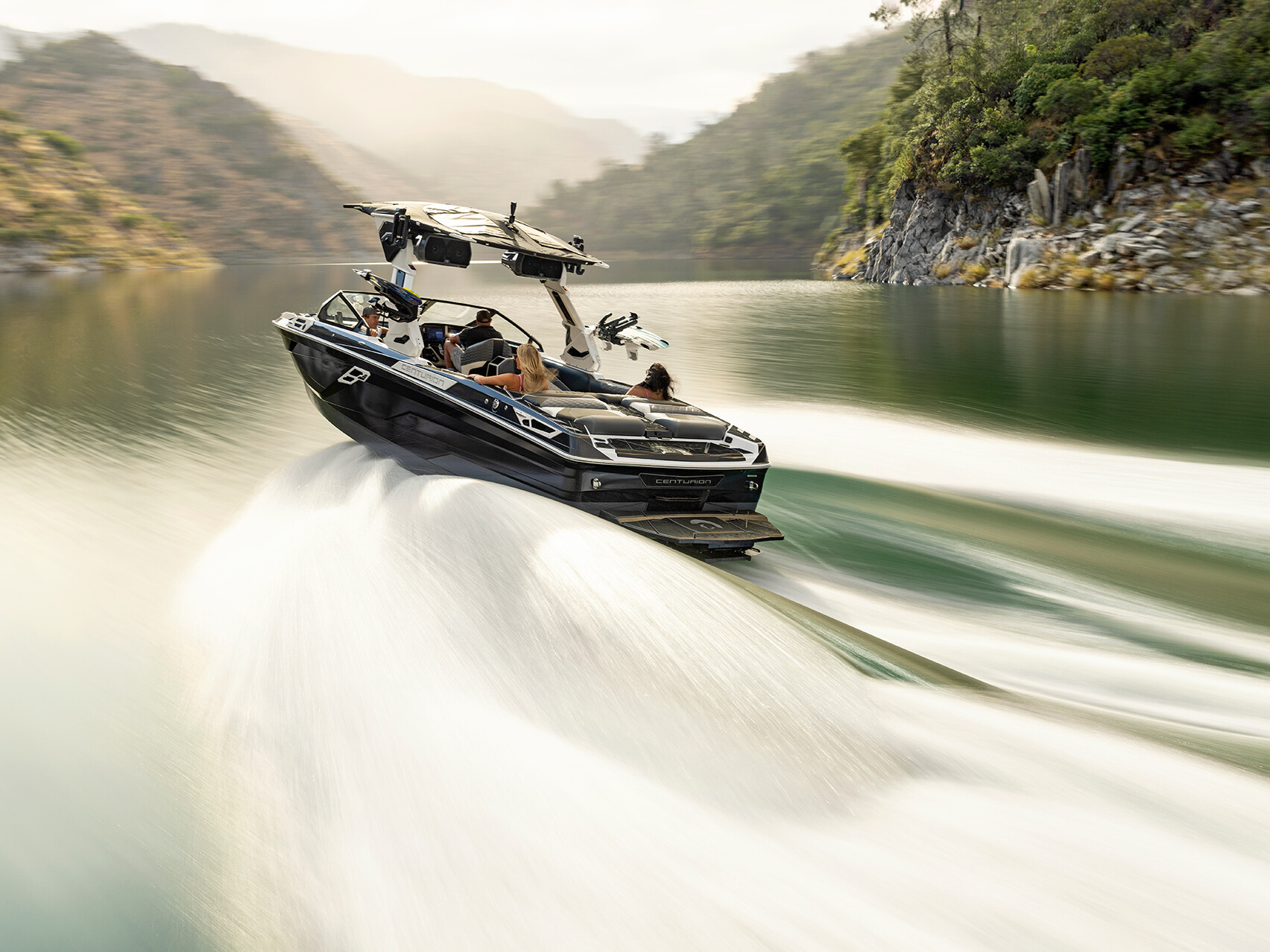 The Centurion Ri Series speedboat with people on board moves swiftly across a calm lake, surrounded by forested hills in the background.