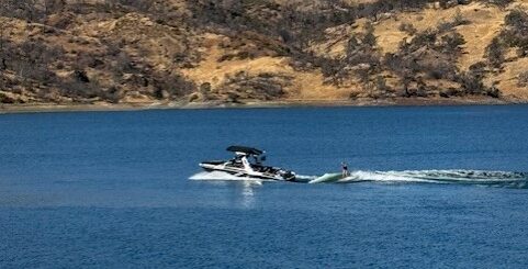 A person wakeboards behind a motorboat on a calm lake during the Cali Comp Festival, with dry, rocky hills and a clear blue sky painting a stunning backdrop.