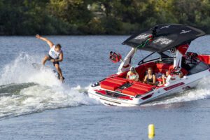 A person wakeboarding performs a jump near a red and white Centurion Boat, full of people onboard, cruising on a calm lake.