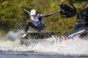 A person wearing a helmet and gloves performs a jump on a wakeboard near the boat, splashing water. Set against vibrant green trees, this exhilarating moment feels straight out of the World Series of Wake Surfing.