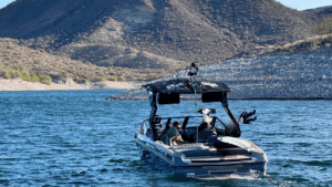 A blue and white Centurion WSWS boat with passengers gracefully navigates a lake surrounded by rocky hills and sparse vegetation, reminiscent of the Wild West, all under a clear sky.