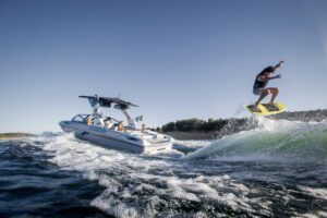 A person is wakeboarding on a wave behind a speedboat on a sunny day, with several people watching from the boat.